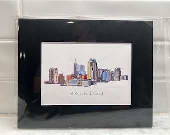 May include: A black framed print with a white background featuring a colorful illustration of the Raleigh, North Carolina skyline. The text "RALEIGH" is printed below the skyline.