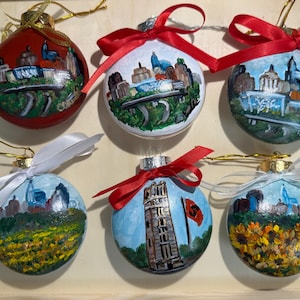 Puede incluir: Seis adornos navideños pintados a mano con paisajes urbanos y monumentos. Cada bola de cristal presenta un diseño diferente, incluyendo un adorno rojo con un paisaje urbano y un adorno azul con la Torre Inclinada de Pisa. Los adornos están adornados con cintas.