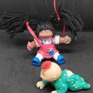 Op de afbeelding: Twee Cabbage Patch Kids poppen. Eén pop heeft zwart haar in staartjes en draagt een roze en blauwe stippenjurk. De andere pop heeft rood haar en draagt een groene en gele stippenpyjama.