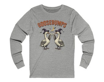 May include: A heather grey long-sleeve t-shirt featuring a graphic of two white geese wearing purple witch hats and performing a fist bump. Above the geese, the text GOOSEBUMPS is printed in an orange, dripping font with a crescent moon and bats.