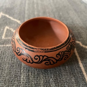 May include: A small, round, terracotta-colored clay bowl featuring a matte finish. The exterior is decorated with a continuous black painted band around the upper section, showcasing a detailed, repeating scrollwork pattern in black ink.