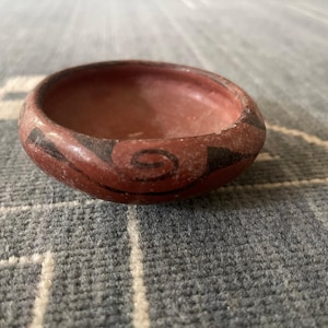 May include: A small, round, terracotta-colored ceramic bowl featuring a smooth, matte finish. The exterior is decorated with hand-painted black spiral and geometric patterns that wrap around the curved sides of the vessel.