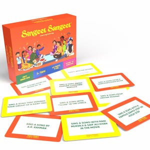 May include: A board game box, orange in colour, labelled "Singeet Sangeet" with illustrations of people singing. Several yellow and orange cards with song prompts are scattered around the box. The game is for 2+ teams.
