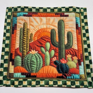 May include: A quilted square featuring a desert landscape with cacti, mountains, and a setting sun. The design uses shades of green, orange, and brown. The border is a green and white checkered pattern.