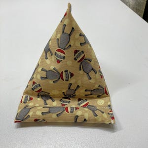 May include: A triangular fabric phone stand featuring a repeating pattern of sock monkey characters in gray, red, and white on a tan background. The stand is folded and sewn, with a small, pointed top.