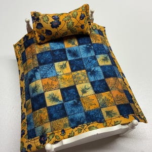 May include: Miniature bed with a white frame, featuring a patchwork quilt in shades of blue, yellow, and orange. The quilt and matching pillow are adorned with floral patterns. A charming dollhouse accessory.