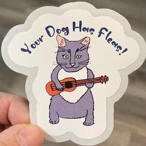 May include: A sticker featuring a cartoon cat playing a ukulele. The cat is purple with a white bib and orange instrument. Text above reads "Your Dog Has Fleas!" in blue, with a light gray cloud-shaped border.