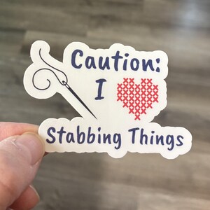 May include: White sticker with a blue needle and thread, a red cross-stitch heart, and the text "Caution: I ♥ Stabbing Things".