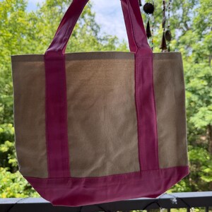 May include: A beige canvas tote bag featuring bright pink fabric accents. The bag includes two vertical pink straps that extend from the base to form the handles, and a solid pink horizontal band along the bottom edge of the rectangular body.