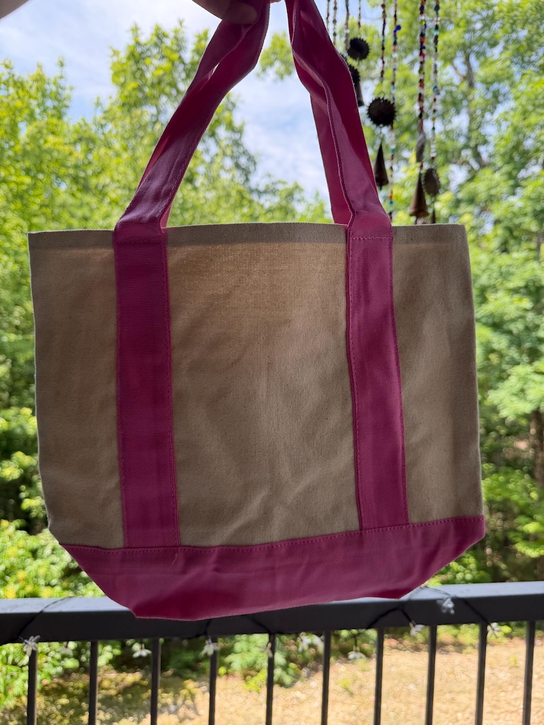 May include: A beige canvas tote bag featuring bright pink fabric accents. The bag includes two vertical pink straps that extend from the base to form the handles, and a solid pink horizontal band along the bottom edge of the rectangular body.