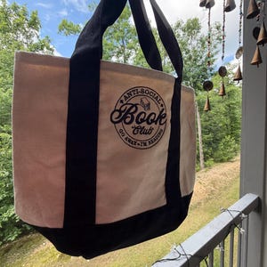May include: A beige canvas tote bag with black handles and a black base. The front features a circular black graphic with the text "ANTI-SOCIAL BOOK CLUB" and "GO AWAY I'M READING" surrounding a small illustration of a stack of books.