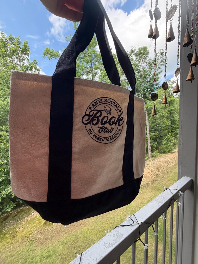 May include: A beige canvas tote bag with black handles and a black base. The front features a circular black graphic with the text "ANTI-SOCIAL BOOK CLUB" and "GO AWAY I'M READING" surrounding a small illustration of a stack of books.