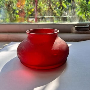 May include: A red glass jar with a decorative design. The jar has a round body and a wide mouth.