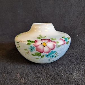 Puede incluir: Un jarrón de vidrio blanco con un diseño floral azul y blanco. El jarrón tiene un cuerpo redondo y una abertura ancha. Las flores están pintadas en rosa, azul y verde.