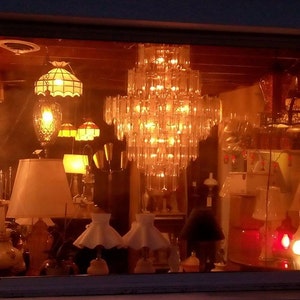 May include: A display of various lamps and light fixtures in a shop window. The lamps are made of glass, metal, and fabric. The light fixtures are a mix of styles, including chandeliers, pendant lights, and table lamps. The lamps are illuminated, casting a warm glow on the display.