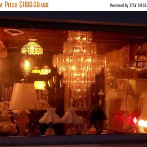 May include: A storefront window display featuring a variety of antique lamps and chandeliers. The display is lit with warm, yellow light, creating a cozy and inviting atmosphere. The lamps are made of glass and metal, and they have a variety of styles, including traditional, modern, and vintage.