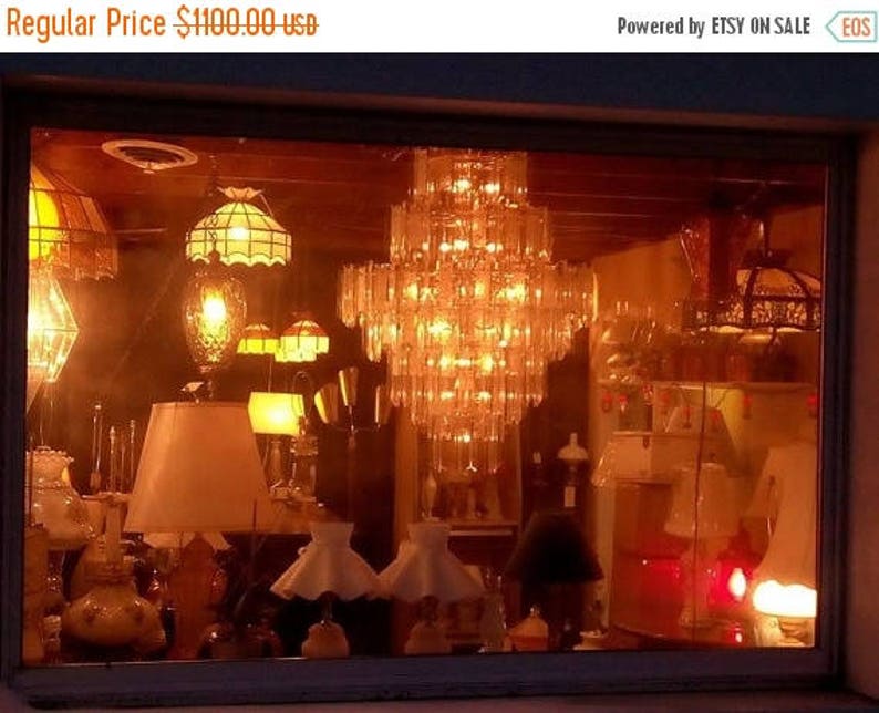 May include: A storefront window display featuring a variety of antique lamps and chandeliers. The display is lit with warm, yellow light, creating a cozy and inviting atmosphere. The lamps are made of glass and metal, and they have a variety of styles, including traditional, modern, and vintage.