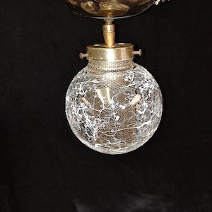Puede incluir: Una lámpara de techo vintage con una pantalla de vidrio esférica. La pantalla es transparente con un diseño de salpicaduras blancas. La lámpara tiene una capota de metal color latón y una banda de metal perforada.