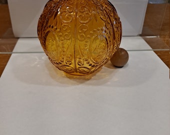May include: A round, amber-colored glass vase with a decorative, embossed pattern. The vase has a smooth, rounded bottom and a slightly flared opening.