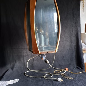 May include: A mid-century modern pendant light fixture with a wood frame and clear glass panels. The light has a silver finish and three light bulbs. The fixture is hanging from a black cord with a plug.