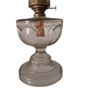 Puede incluir: Una lámpara de aceite de vidrio transparente con un quemador de latón y una base decorativa. La base tiene un diseño de clave griega.