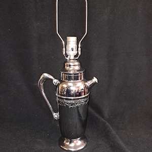 May include: A silver cocktail shaker lamp with a decorative design around the middle. The lamp has a simple metal shade.