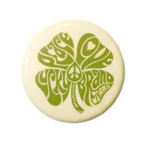 May include: A circular, cream-coloured button featuring a green four-leaf clover design. The clover incorporates a peace symbol and the words "Lucky Brand Jeans". The button has a glossy finish.