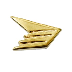 May include: A gold-coloured metal pin in the shape of a stylised wing or arrow. The pin has a smooth, polished surface and a three-dimensional design with layered details. The pin is set against a white background.