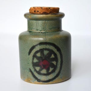 May include: A cylindrical ceramic jar with a mottled sage green glaze and a cork stopper. The front features a hand-painted black circular motif resembling a wheel with a small red centre. The jar has a smooth, glossy finish and a wide rim.