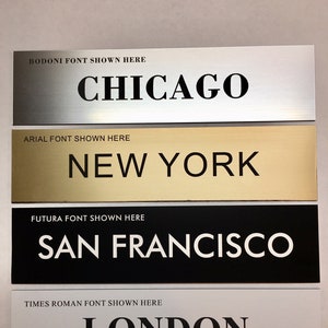 May include: Four metal plaques with different fonts and city names. The top plaque is silver with the text "BODONI FONT SHOWN HERE CHICAGO". The second plaque is gold with the text "ARIAL FONT SHOWN HERE NEW YORK". The third plaque is black with the text "FUTURA FONT SHOWN HERE SAN FRANCISCO". The bottom plaque is silver with the text "TIMES ROMAN FONT SHOWN HERE LONDON".