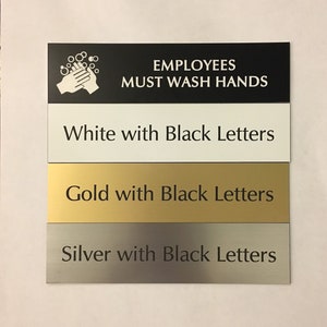 May include: Four signs with different colored backgrounds and black lettering. The top sign is black with white lettering that reads "Employees Must Wash Hands". The sign also has a white icon of two hands washing with soap and water. The second sign is white with black lettering that reads "White with Black Letters". The third sign is gold with black lettering that reads "Gold with Black Letters". The fourth sign is silver with black lettering that reads "Silver with Black Letters".