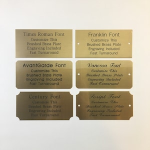 May include: Six gold-colored metal plaques with different font styles. Each plaque has the font name, "Customize This", "Brushed Brass Plate", "Engraving Included", and "Fast Turnaround" printed on it.