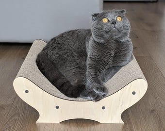 May include: A gray Scottish Fold cat lounges on a beige and wood-toned cat scratcher. The cat has yellow eyes and is looking upwards. The scratcher has a curved design with a corrugated surface and wooden sides, with black dot accents.