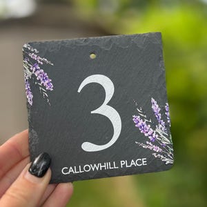 May include: A square, black slate house number sign featuring the number 3 in white. The sign is decorated with lavender floral accents and the text "CALLOWHILL PLACE" in white. A hole is drilled at the top for hanging.