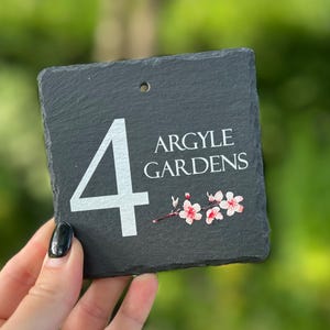 May include: A square, black slate house number sign displaying the number "4" in white, the words "ARGYLE GARDENS", and a floral design. The sign has a small hole at the top for hanging.
