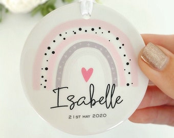 Op de afbeelding: Een witte keramische ornament met een roze en grijze regenboog, zwarte stippen en een roze hart. De ornament heeft de naam "Isabelle" en de datum "21 mei 2020" erop.