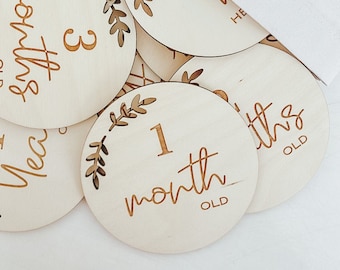 Può includere: Un set di dischi in legno per le tappe del bambino con un design a foglia e lettere dorate. I dischi presentano i numeri da 1 a 12, con le parole "mese" o "anno" scritte sotto ogni numero. I dischi sono disposti in una pila, con una borsa di stoffa bianca visibile sullo sfondo.
