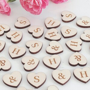 May include: Wooden heart-shaped pieces with the letters H and S and the ampersand symbol. The pieces are scattered on a white surface with pink flowers in the background.