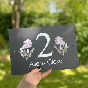 May include: A rectangular black slate house number sign with the number "2" in white, floral accents, and the text "Allens Close" in white. The sign is held up against a blurred green background.