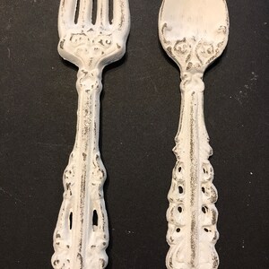 May include: A white metal fork and spoon with ornate designs. The utensils have a distressed white finish with visible gray accents.