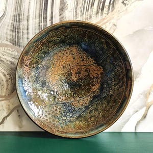 May include: A round ceramic bowl featuring a textured surface with a repeating pattern of small, raised circles. The glaze displays a gradient of metallic bronze, deep blue, and earthy brown tones, creating a reflective and variegated finish.