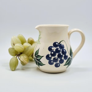 May include: Cream-coloured ceramic jug with a painted design of blue grapes and green leaves. The jug has a handle and a spout. A bunch of green grapes is next to the jug.