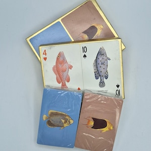 May include: A collection of playing cards featuring fish illustrations. Two decks are wrapped in clear plastic, one blue and one tan. Behind them, two loose cards show a four of hearts with a red fish and a ten of spades with a spotted fish.