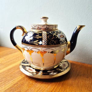 May include: A decorative porcelain teapot and saucer set. The teapot has a cream base with gold and orange floral patterns, a navy blue band with gold detailing, and a gold-edged lid and handle. The saucer complements the teapot's design with similar patterns.