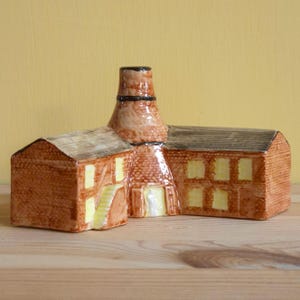 May include: A ceramic building figurine with a chimney, featuring a brown brick exterior, yellow window accents, and a dark roof. The figurine has a detailed design, with a staircase and door.