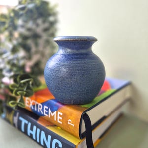 May include: A small, blue ceramic vase with a textured surface and a narrow neck. The vase is sitting on top of two books with colourful covers. The vase is a decorative item.