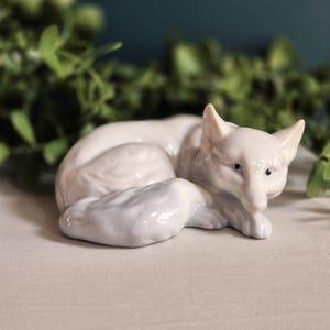 May include: A white ceramic mug with a black handle and the text "But First, Coffee" in a bold, black font. The mug is set against a blurred background, suggesting a cosy setting.