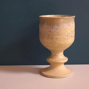 May include: A ceramic goblet with a speckled beige and tan finish. The vessel features a wide, rounded bowl that transitions into a narrow stem with a prominent central ring, resting on a circular base. The glaze shows subtle lavender hues.