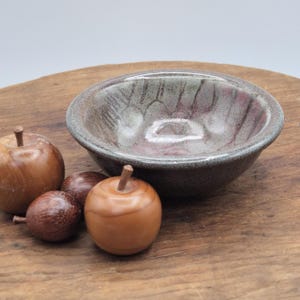 May include: A round ceramic bowl with a mottled grey and reddish-brown glaze featuring a subtle ribbed texture on the interior, accompanied by three smooth, polished wooden apple-shaped ornaments with small brown stems.