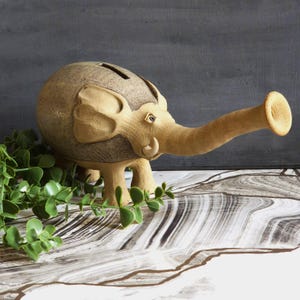 May include: A ceramic elephant-shaped money box in tan and brown hues. The elephant features large ears, a long trunk, and a slot on its back for coins. It rests on a marbled surface with green foliage.